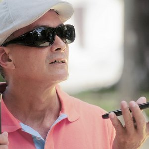 Mann mit schwarzer Brille, Blindenstock kommuniziert über sein Smartphone